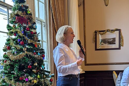 Susan Murray giving a speech, holding a microphone in front of a decorated Christmas tree.